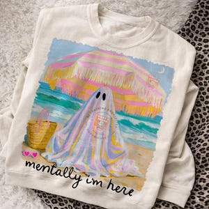 May include: A cream-colored crewneck sweatshirt featuring a graphic of a ghost sitting under a pink and yellow striped beach umbrella on a sandy beach with a straw tote bag. The text "mentally i'm here" is printed in black cursive below the graphic.