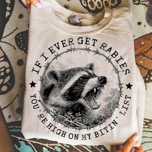 Puede incluir: Sudadera color crema con una ilustración en blanco y negro de un mapache gruñendo dentro de un círculo de alambre de púas. El texto dice: "IF I EVER GET RABIES, YOU'RE HIGH ON MY BITIN' LIST."