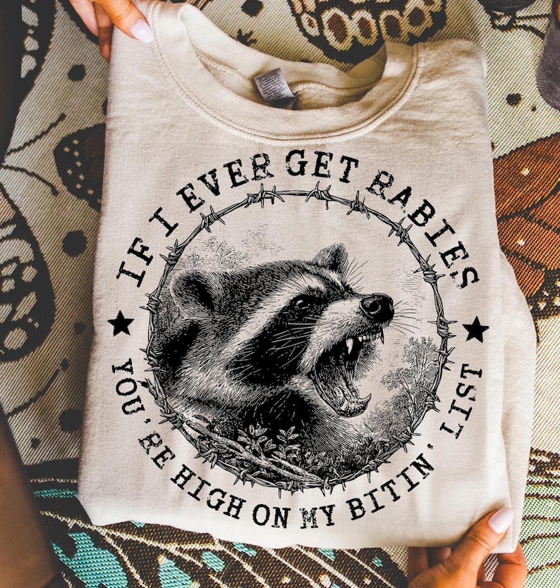 May include: A cream-colored sweatshirt featuring a black and white illustration of a snarling raccoon within a barbed wire circle. The text reads "IF I EVER GET RABIES, YOU'RE HIGH ON MY BITIN' LIST."