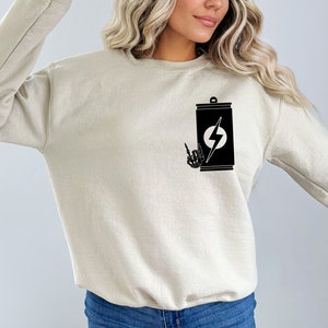 May include: A beige sweatshirt with a black graphic of a can with a lightning bolt and a skeleton hand giving the rock and roll sign.
