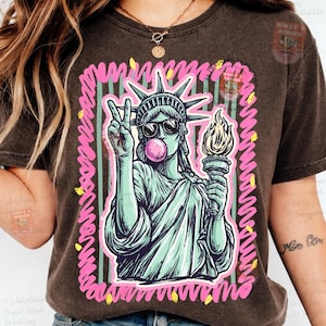 Puede incluir: Camiseta marr&oacute;n oscuro con un gr&aacute;fico de la Estatua de la Libertad con gafas de sol y soplando una burbuja de chicle. La estatua sostiene una antorcha y hace el signo de la paz. El dise&ntilde;o est&aacute; enmarcado con l&iacute;neas rosas y amarillas.