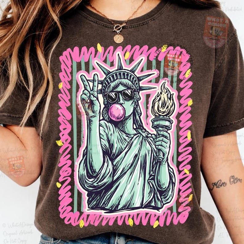 Puede incluir: Camiseta marr&oacute;n oscuro con un gr&aacute;fico de la Estatua de la Libertad con gafas de sol y soplando una burbuja de chicle. La estatua sostiene una antorcha y hace el signo de la paz. El dise&ntilde;o est&aacute; enmarcado con l&iacute;neas rosas y amarillas.