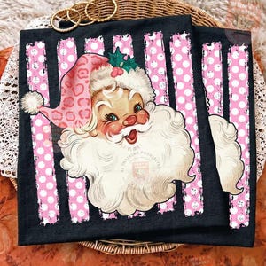 Pode incluir: Uma t-shirt preta com um gr&aacute;fico do Pai Natal a usar um gorro cor-de-rosa com padr&atilde;o de leopardo, um pompom branco e folhas de azevinho verdes. O fundo do gr&aacute;fico consiste em riscas verticais cor-de-rosa com bolas brancas.