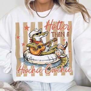 May include: White sweatshirt with a graphic of an alligator wearing a cowboy hat and boots, playing a guitar while floating on an inner tube. The text reads "Hotter Than A Hoochie Coochie" in orange script.