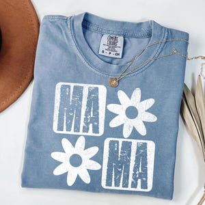 Peut inclure: Un t-shirt bleu clair avec un motif graphique blanc qui dit "Mama" avec une marguerite entre les mots.