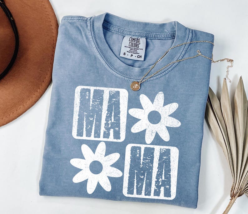 Peut inclure: Un t-shirt bleu clair avec un motif graphique blanc qui dit "Mama" avec une marguerite entre les mots.