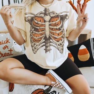 May include: Cream-colored t-shirt featuring a skeletal rib cage design with three orange jack-o'-lanterns inside. The shirt is part of a Halloween-themed collection. The person is wearing black shorts and brown and white checkered shoes.