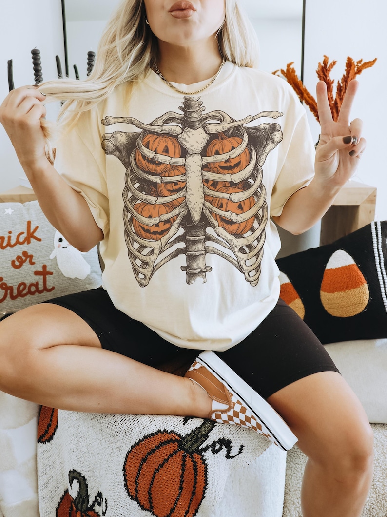 May include: Cream-colored t-shirt featuring a skeletal rib cage design with three orange jack-o'-lanterns inside. The shirt is part of a Halloween-themed collection. The person is wearing black shorts and brown and white checkered shoes.