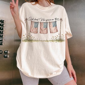 May include: Cream-colored t-shirt featuring a graphic of three American flags hanging on a clothesline, with the text "God shed His grace on thee." The flags have red and white stripes and blue stars. The shirt has a relaxed fit.