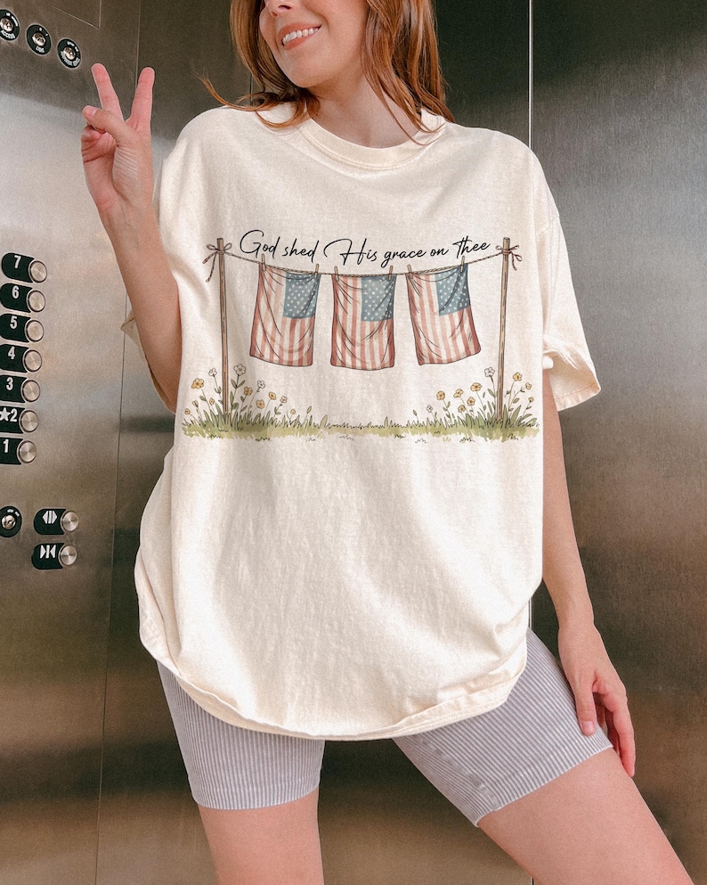 May include: Cream-colored t-shirt featuring a graphic of three American flags hanging on a clothesline, with the text "God shed His grace on thee." The flags have red and white stripes and blue stars. The shirt has a relaxed fit.