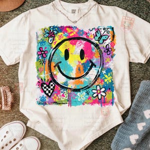 May include: Cream-colored t-shirt featuring a large, colorful smiley face design. The graphic includes a smiley face, flowers, butterflies, and a checkered heart. The shirt has a crew neck and short sleeves.