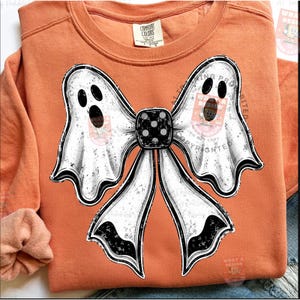 Op de afbeelding: Een oranje sweatshirt met lange mouwen en een grote afbeelding van een strik gemaakt van twee witte geesten met zwarte ogen en monden. Het midden van de strik is een zwarte dobbelsteen met witte stippen. Het kledingstuk heeft een Comfort Colors label.