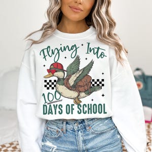 May include: White sweatshirt with a green duck wearing sunglasses and a red hat flying with a backpack. The text "Flying Into 100 Days of School" is printed on the sweatshirt.