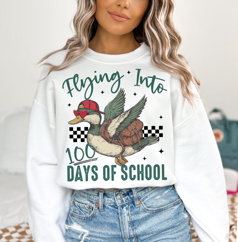 May include: White sweatshirt with a green duck wearing sunglasses and a red hat flying with a backpack. The text "Flying Into 100 Days of School" is printed on the sweatshirt.