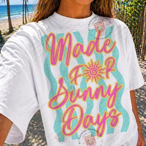 May include: White t-shirt with the text "Made For Sunny Days" in pink and yellow script, with a sun graphic. The text is layered over wavy turquoise stripes. The shirt has short sleeves.