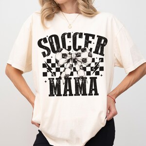 Puede incluir: Una camiseta blanca con un dise&ntilde;o gr&aacute;fico negro que dice "Soccer Mama" con un patr&oacute;n de cuadros blanco y negro y un lazo de bal&oacute;n de f&uacute;tbol.