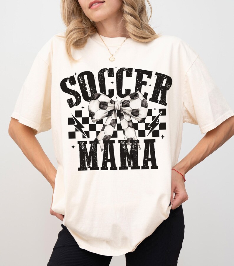 Puede incluir: Una camiseta blanca con un dise&ntilde;o gr&aacute;fico negro que dice "Soccer Mama" con un patr&oacute;n de cuadros blanco y negro y un lazo de bal&oacute;n de f&uacute;tbol.
