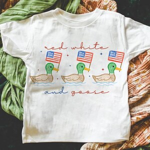 May include: White t-shirt with a patriotic design. The shirt features three ducks holding American flags, with the words "red white and goose" in red and blue script. The ducks have green heads, tan bodies, and yellow beaks.