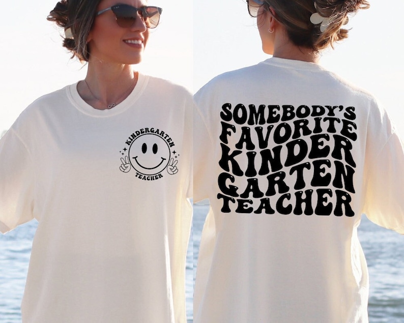 May include: A cream-coloured t-shirt with black text and graphics. The front displays a smiley face with peace signs and the words "Kindergarten Teacher." The back reads "Somebody's Favorite Kindergarten Teacher" in a retro font.