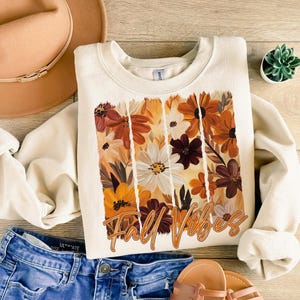 May include: Cream-colored sweatshirt with a floral design in shades of orange, brown, and white. The words "Fall Vibes" are printed in a cursive font. The sweatshirt is paired with blue jeans, a tan hat, and sandals.