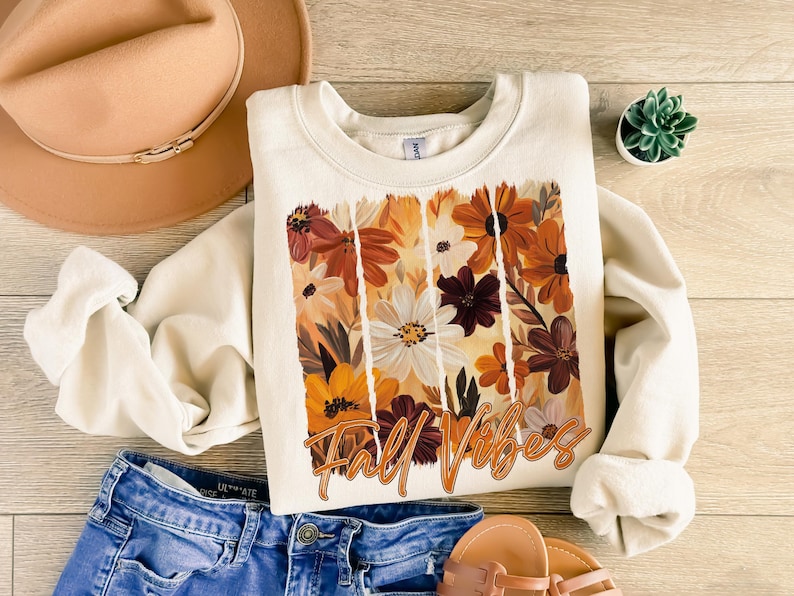 May include: Cream-colored sweatshirt with a floral design in shades of orange, brown, and white. The words "Fall Vibes" are printed in a cursive font. The sweatshirt is paired with blue jeans, a tan hat, and sandals.
