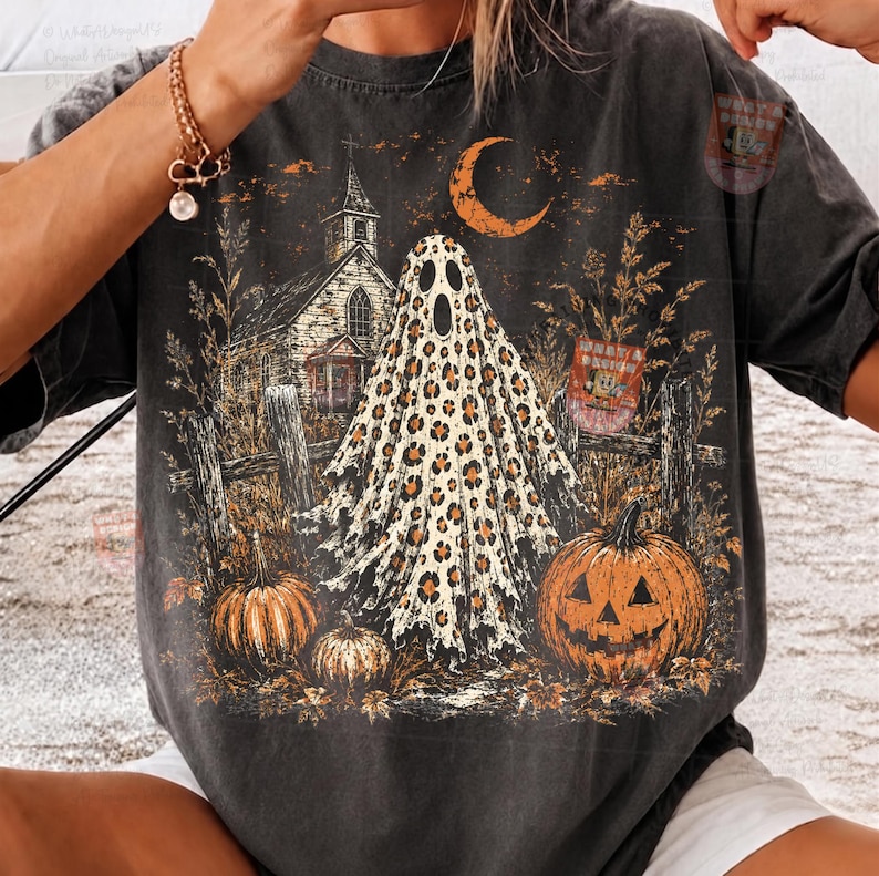 May include: A charcoal gray short-sleeve t-shirt featuring a graphic print of a ghost with a leopard print pattern, a church building, a crescent moon, and two pumpkins. The design includes tall grass, a wooden fence, and a small rectangular sign.