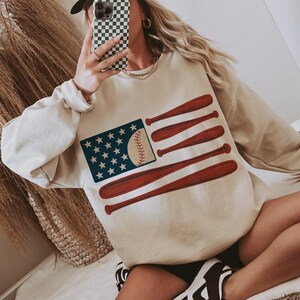 May include: Beige sweatshirt featuring a baseball-themed American flag design. The flag includes a baseball in place of the stripes and bats as the stripes. The image also shows a white baseball cap with the text "Malibu Cowboy".