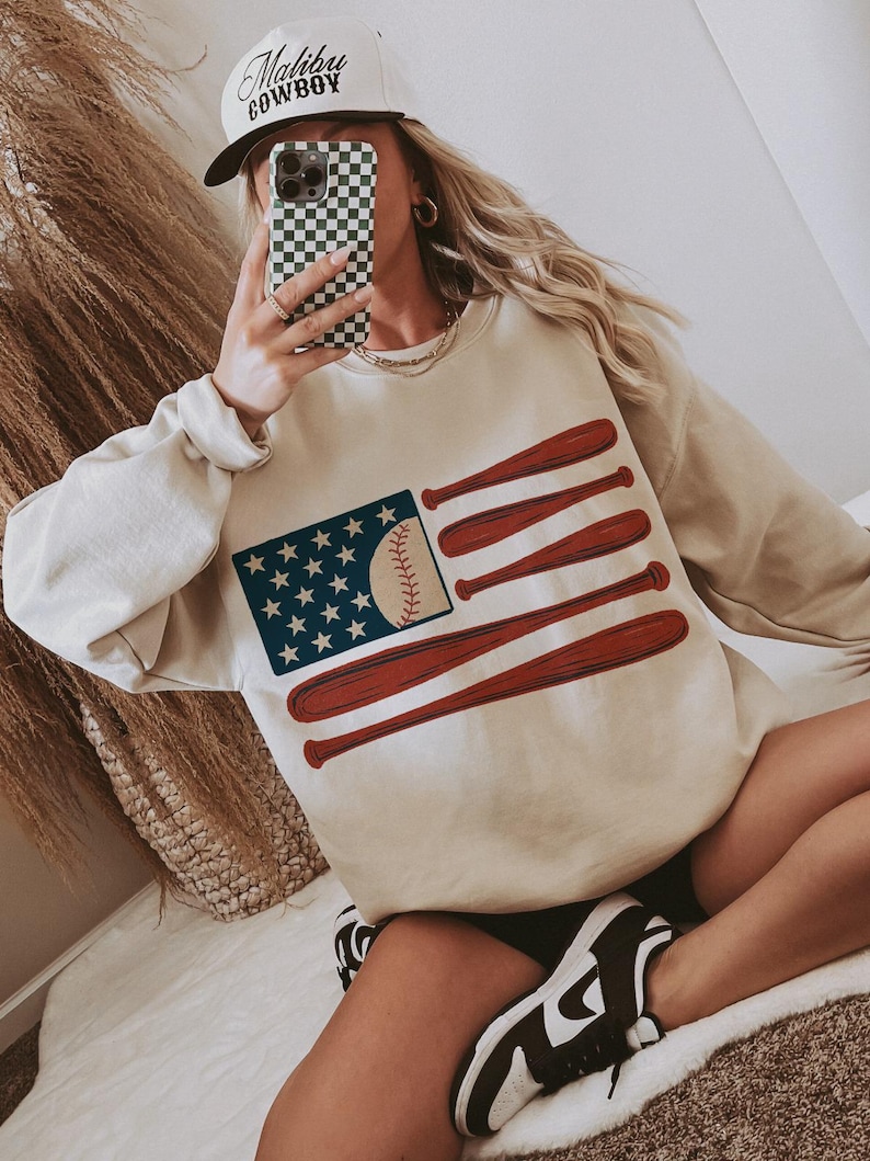 May include: Beige sweatshirt featuring a baseball-themed American flag design. The flag includes a baseball in place of the stripes and bats as the stripes. The image also shows a white baseball cap with the text "Malibu Cowboy".