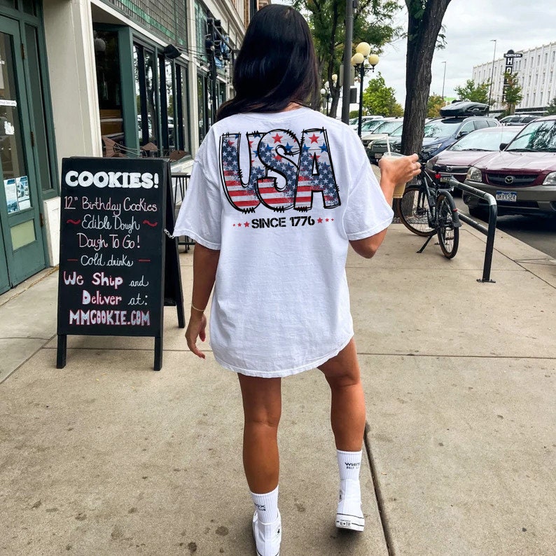 May include: White t-shirt with a distressed American flag design that says "USA" and "Since 1776" in black text.