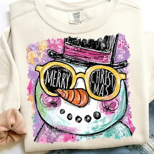 May include: Cream-colored sweatshirt featuring a whimsical snowman design. The snowman wears yellow sunglasses with the words "MERRY CHRISTMAS" and a black top hat with a pink band. The design includes a carrot nose and a colorful background with snowflakes.