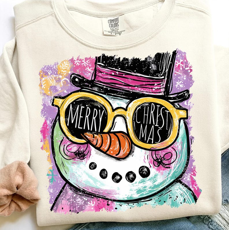 May include: Cream-colored sweatshirt featuring a whimsical snowman design. The snowman wears yellow sunglasses with the words "MERRY CHRISTMAS" and a black top hat with a pink band. The design includes a carrot nose and a colorful background with snowflakes.
