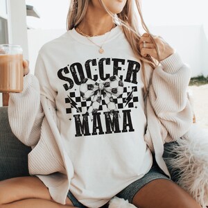 Puede incluir: Sudadera blanca con texto negro que dice "Soccer Mama" con un lazo a cuadros blanco y negro y balones de f&uacute;tbol en el centro.