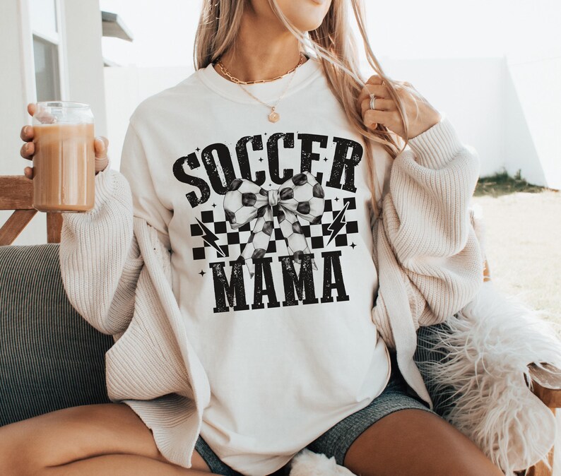 Puede incluir: Sudadera blanca con texto negro que dice "Soccer Mama" con un lazo a cuadros blanco y negro y balones de f&uacute;tbol en el centro.