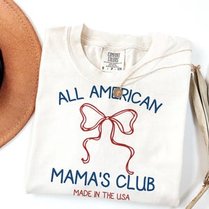 May include: Cream-colored t-shirt with the text "ALL AMERICAN MAMA'S CLUB" in blue, featuring a red bow graphic. The shirt is labeled "MADE IN THE USA". A gold necklace and a brown hat are partially visible.