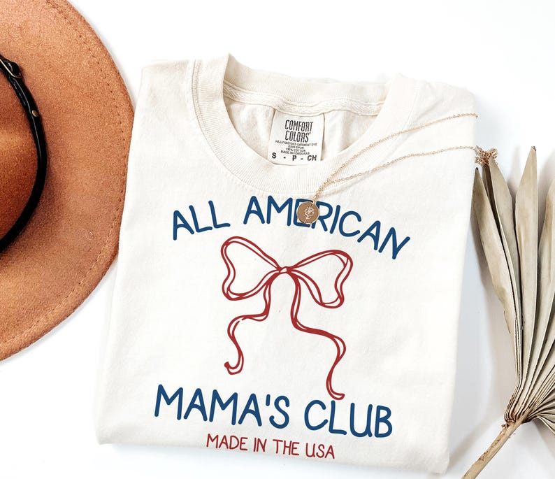 May include: Cream-colored t-shirt with the text "ALL AMERICAN MAMA'S CLUB" in blue, featuring a red bow graphic. The shirt is labeled "MADE IN THE USA". A gold necklace and a brown hat are partially visible.
