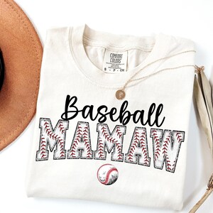 May include: A beige t-shirt with the text "Baseball Mamaw" in black lettering. The letters are outlined with a red and white baseball stitch design. A red and white baseball is centred below the text.