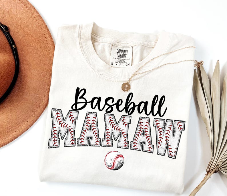 May include: A beige t-shirt with the text "Baseball Mamaw" in black lettering. The letters are outlined with a red and white baseball stitch design. A red and white baseball is centred below the text.