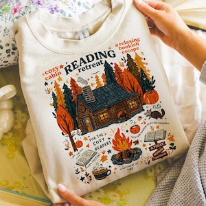 Pode incluir: Camisola creme com um gr&aacute;fico colorido de uma cabana na floresta, uma fogueira e as palavras "Reading Retreat". O design inclui ab&oacute;boras, &aacute;rvores e as frases "cozy cabin" e "a relaxing bookish escape."