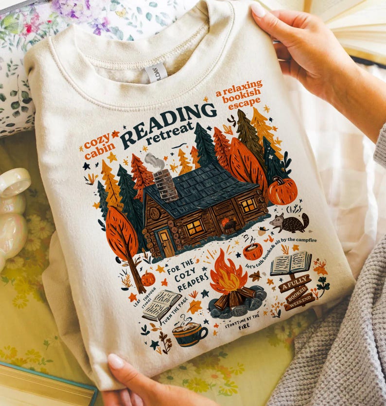 Pode incluir: Camisola creme com um gr&aacute;fico colorido de uma cabana na floresta, uma fogueira e as palavras "Reading Retreat". O design inclui ab&oacute;boras, &aacute;rvores e as frases "cozy cabin" e "a relaxing bookish escape."