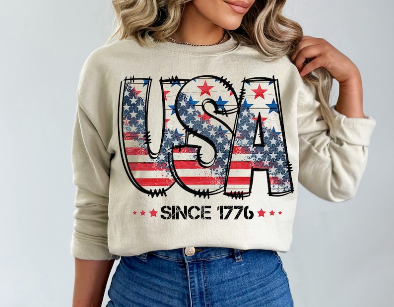 May include: A beige sweatshirt with a graphic print of the word "USA" in red, white, and blue with stars and stripes. The text "Since 1776" is printed below the graphic.