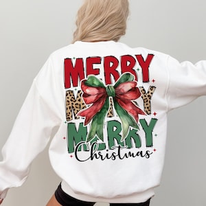 K&ouml;nnte beinhalten: Wei&szlig;er Pullover mit einem roten, gr&uuml;nen und schwarzen Grafikdruck, auf dem "Merry Merry Christmas" mit einer gro&szlig;en gr&uuml;nen und roten Schleife steht.