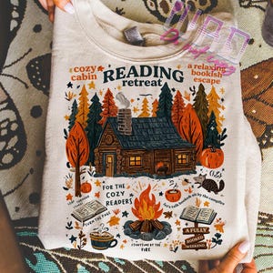 May include: Cream-colored sweatshirt with a colorful graphic of a cabin in the woods, a campfire, and the words "Reading Retreat." The design includes pumpkins, trees, and the phrases "Cozy Cabin" and "A Relaxing Bookish Escape."