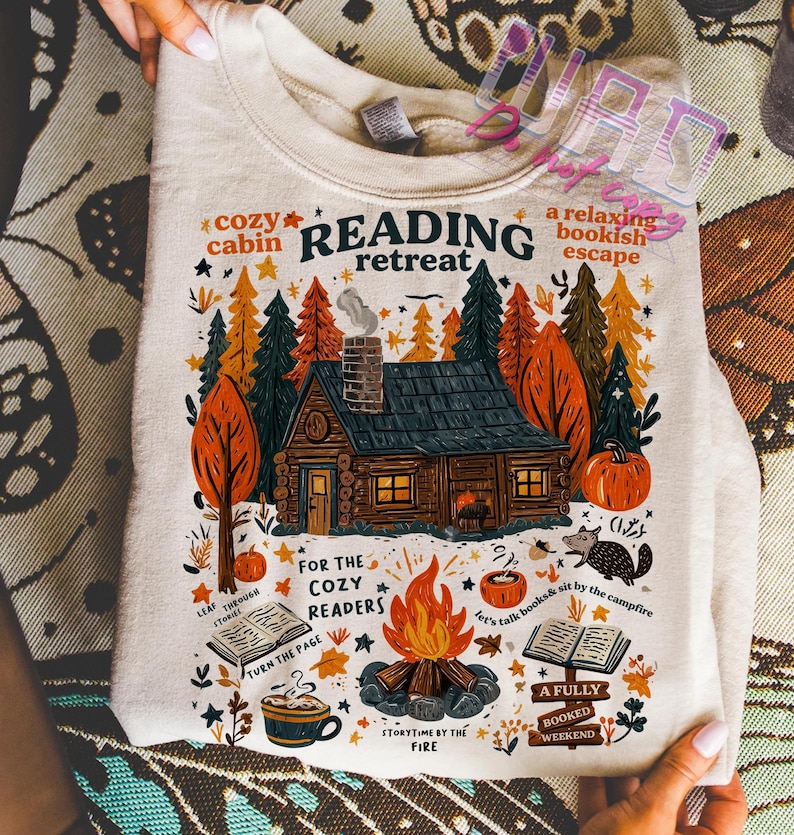 May include: Cream-colored sweatshirt with a colorful graphic of a cabin in the woods, a campfire, and the words "Reading Retreat." The design includes pumpkins, trees, and the phrases "Cozy Cabin" and "A Relaxing Bookish Escape."