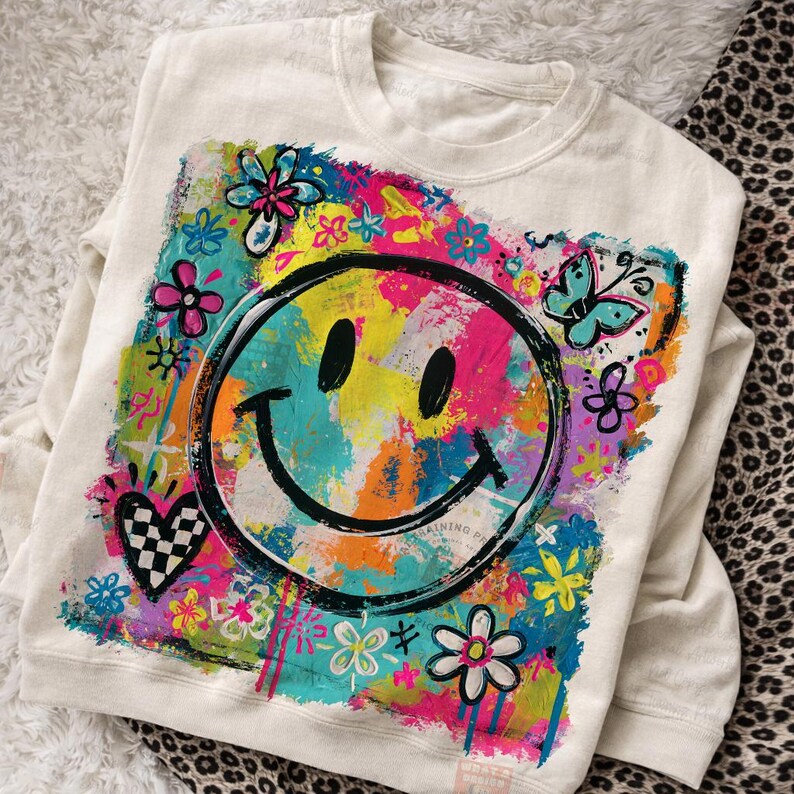 May include: Cream-colored sweatshirt featuring a large, colorful smiley face design. The graphic includes a black outline of a smiley face with a vibrant background of flowers, butterflies, and abstract paint strokes in various colors.