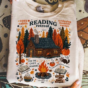 May include: A cream-colored sweatshirt featuring a colorful illustration of a cozy cabin in a forest setting. The design includes a campfire, pumpkins, and text that reads "Reading Retreat" and "Storytime by the Fire."