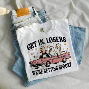 K&ouml;nnte beinhalten: Wei&szlig;es T-Shirt mit dem Text "GET IN, LOSERS WE'RE GETTING SPOOKY" und einer Grafik von Enten in Halloween-Kost&uuml;men in einem rosa Auto. Das T-Shirt liegt auf einer blauen Jeans.