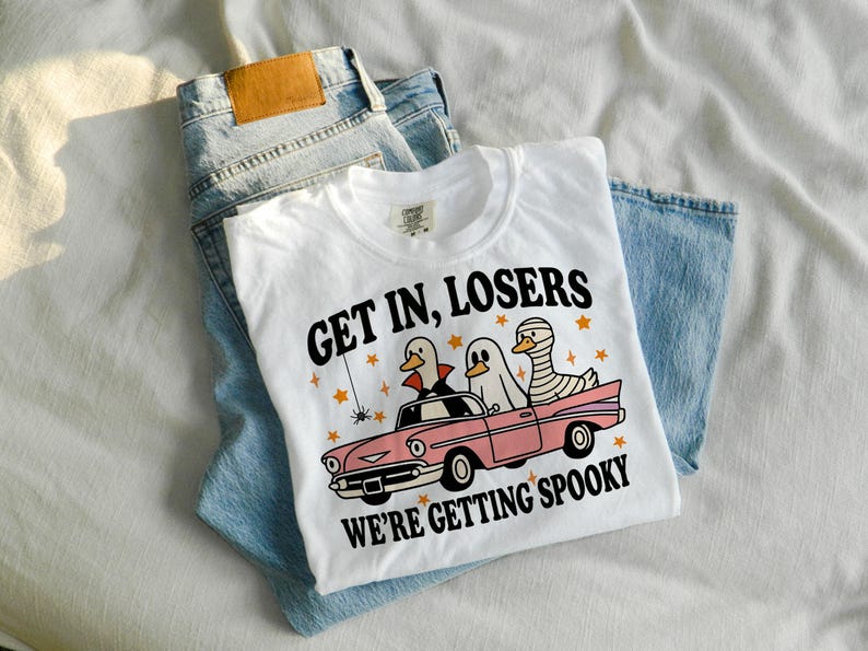K&ouml;nnte beinhalten: Wei&szlig;es T-Shirt mit dem Text "GET IN, LOSERS WE'RE GETTING SPOOKY" und einer Grafik von Enten in Halloween-Kost&uuml;men in einem rosa Auto. Das T-Shirt liegt auf einer blauen Jeans.
