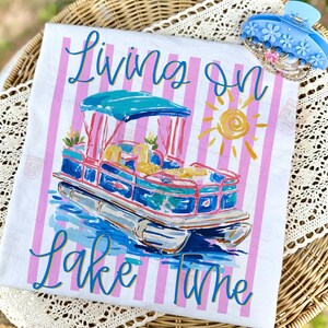 May include: White t-shirt with a colorful illustration of a pontoon boat, the text "Living on Lake Time", and pink vertical stripes. A blue floral hair clip rests on the side. The shirt is displayed on a wicker tray.