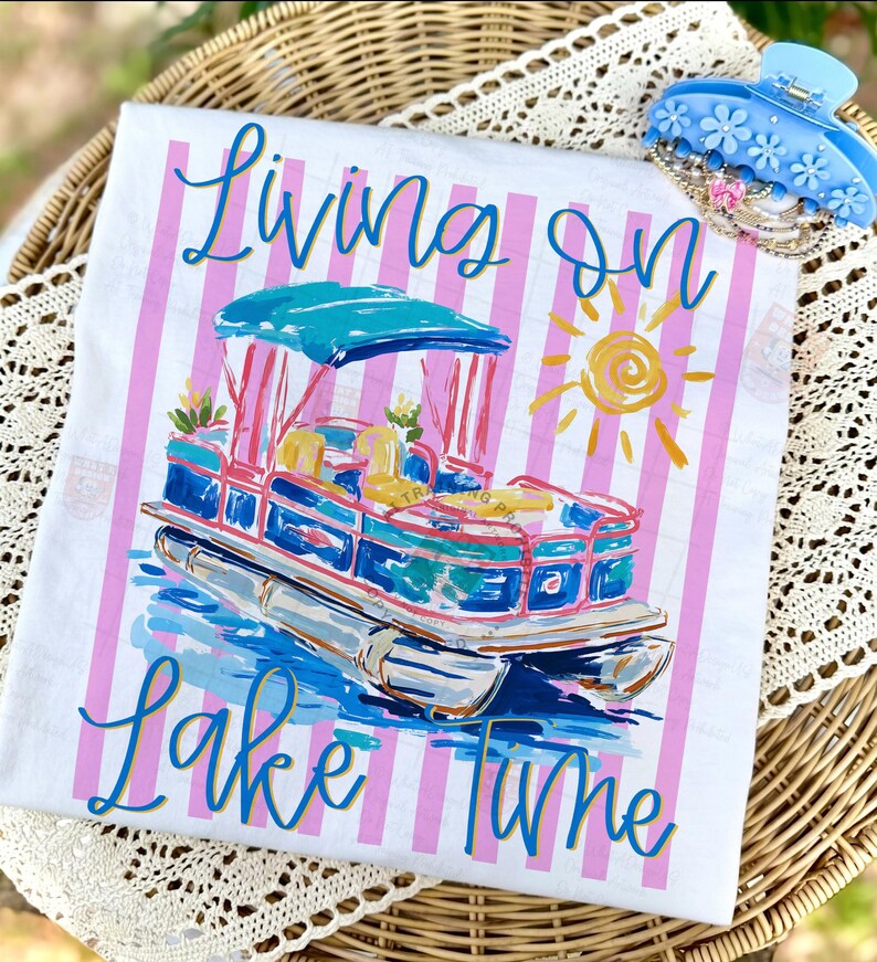 May include: White t-shirt with a colorful illustration of a pontoon boat, the text "Living on Lake Time", and pink vertical stripes. A blue floral hair clip rests on the side. The shirt is displayed on a wicker tray.
