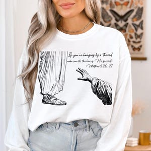 May include: White sweatshirt with a black and white illustration of a hand reaching out. The text on the sweatshirt reads "If you're hanging by a thread make sure it's the hem of His garment. Matthew 9:20-27".